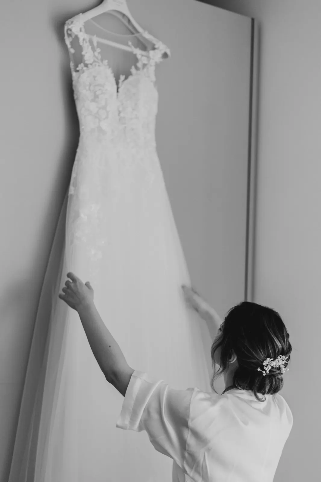 The bride in her robe arranging the lace dress hanging on the wall, in black and white