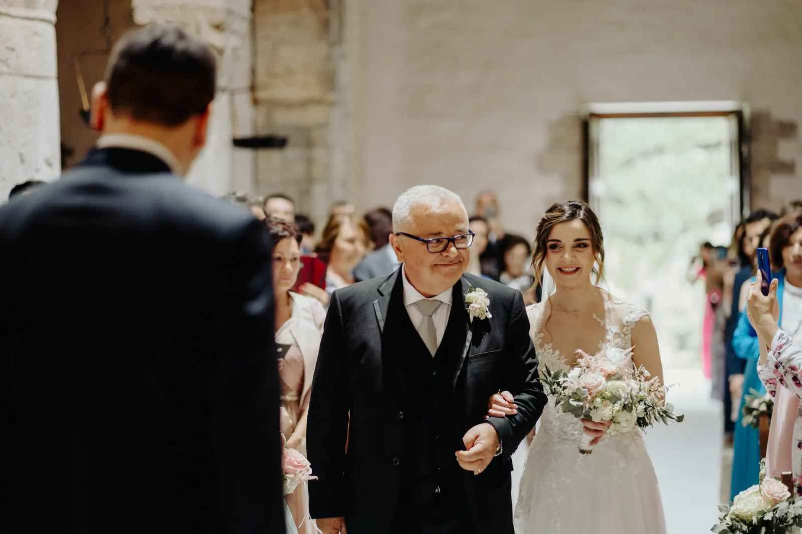 Martina being walked down the aisle, with the groom waiting, seen from behind