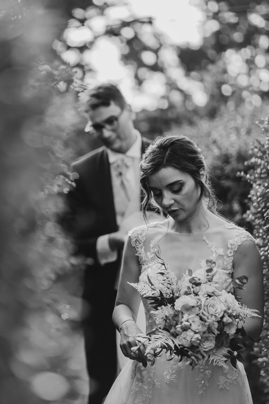 The bride looking at her bouquet with the groom behind her, in black and white