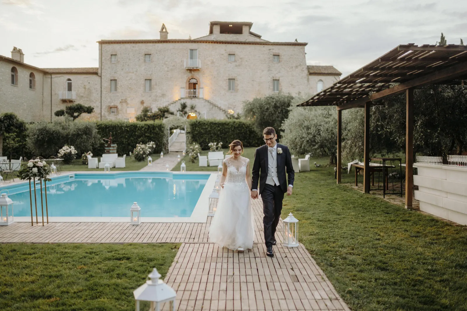 The couple walking hand in hand beside the pool, with Castello di Montignano behind them