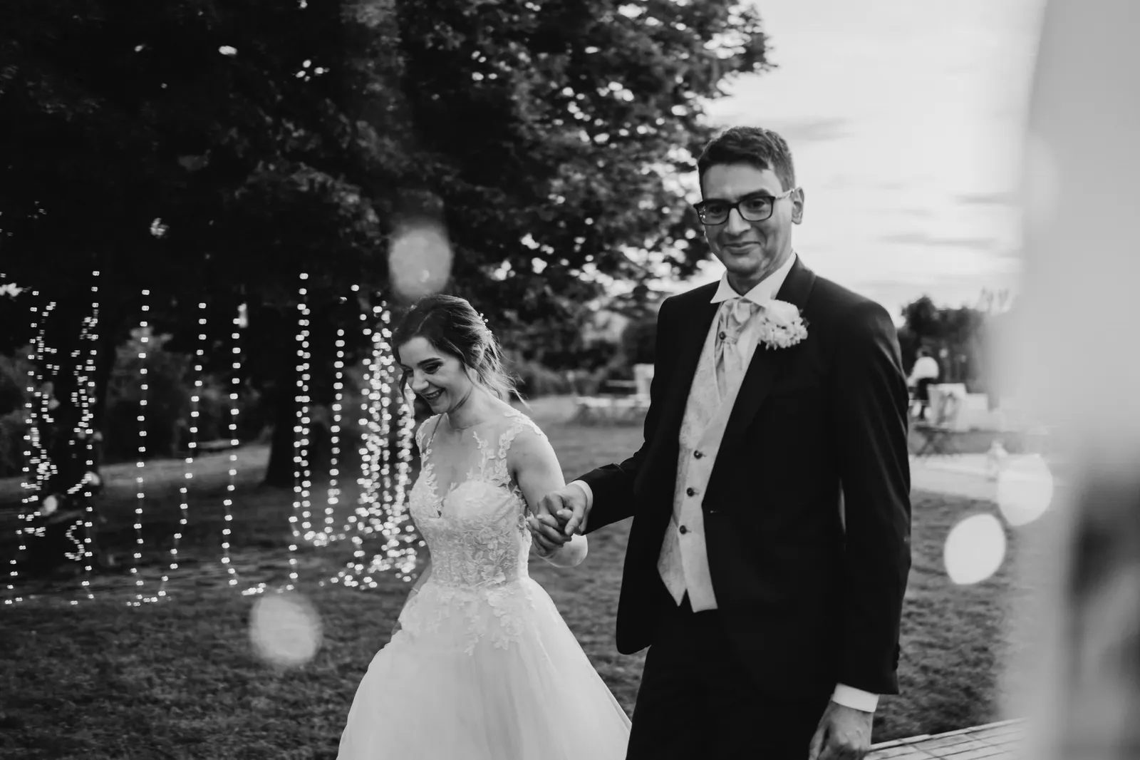 Martina and Daniele arriving at the reception among lights hung from the trees, in black and white