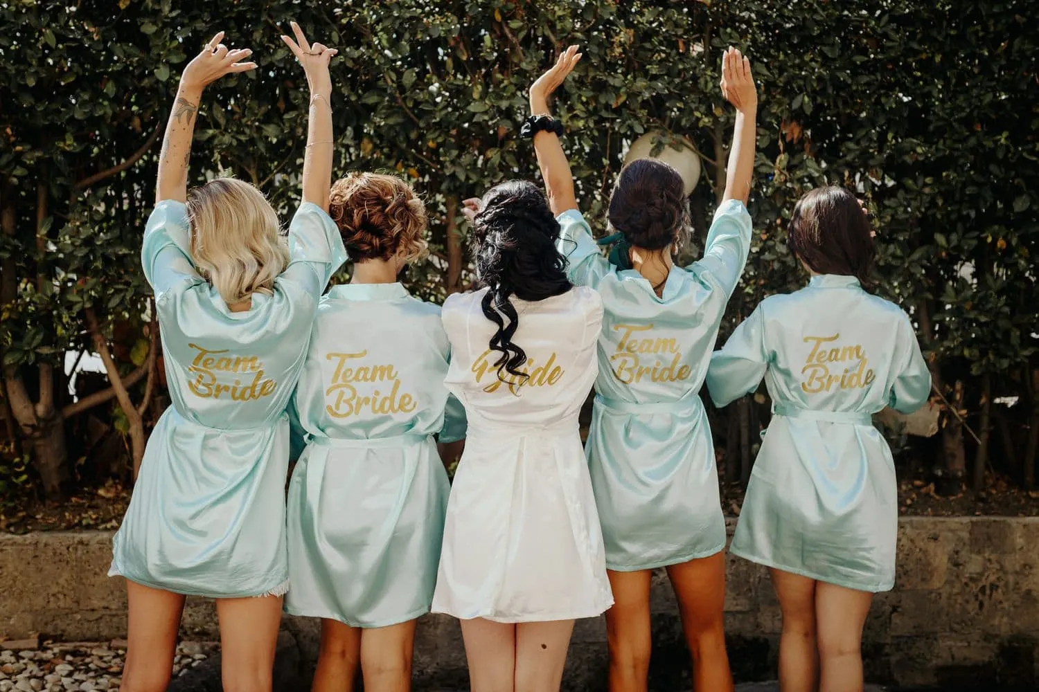 Le amiche di spalle con le vestaglie Team Bride e le braccia alzate