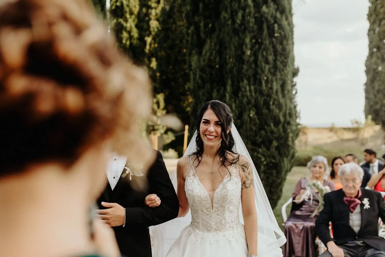 Micaela sorride tra gli invitati, vista oltre la spalla di una damigella