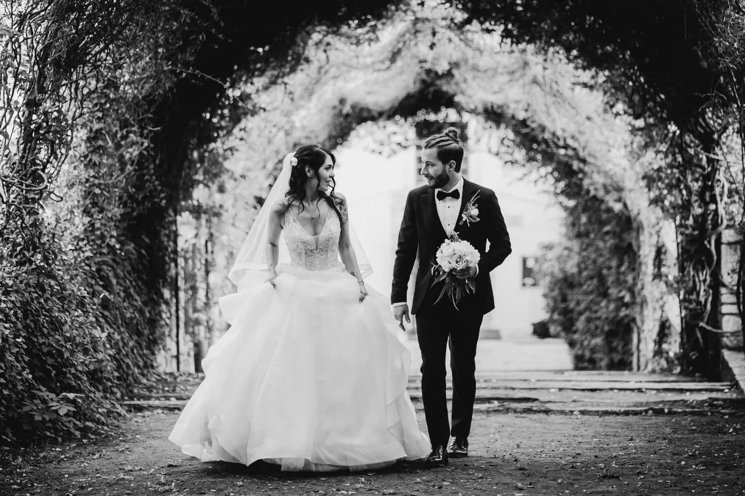 Micaela e Raffaele camminano sotto la galleria di alberi, in bianco e nero