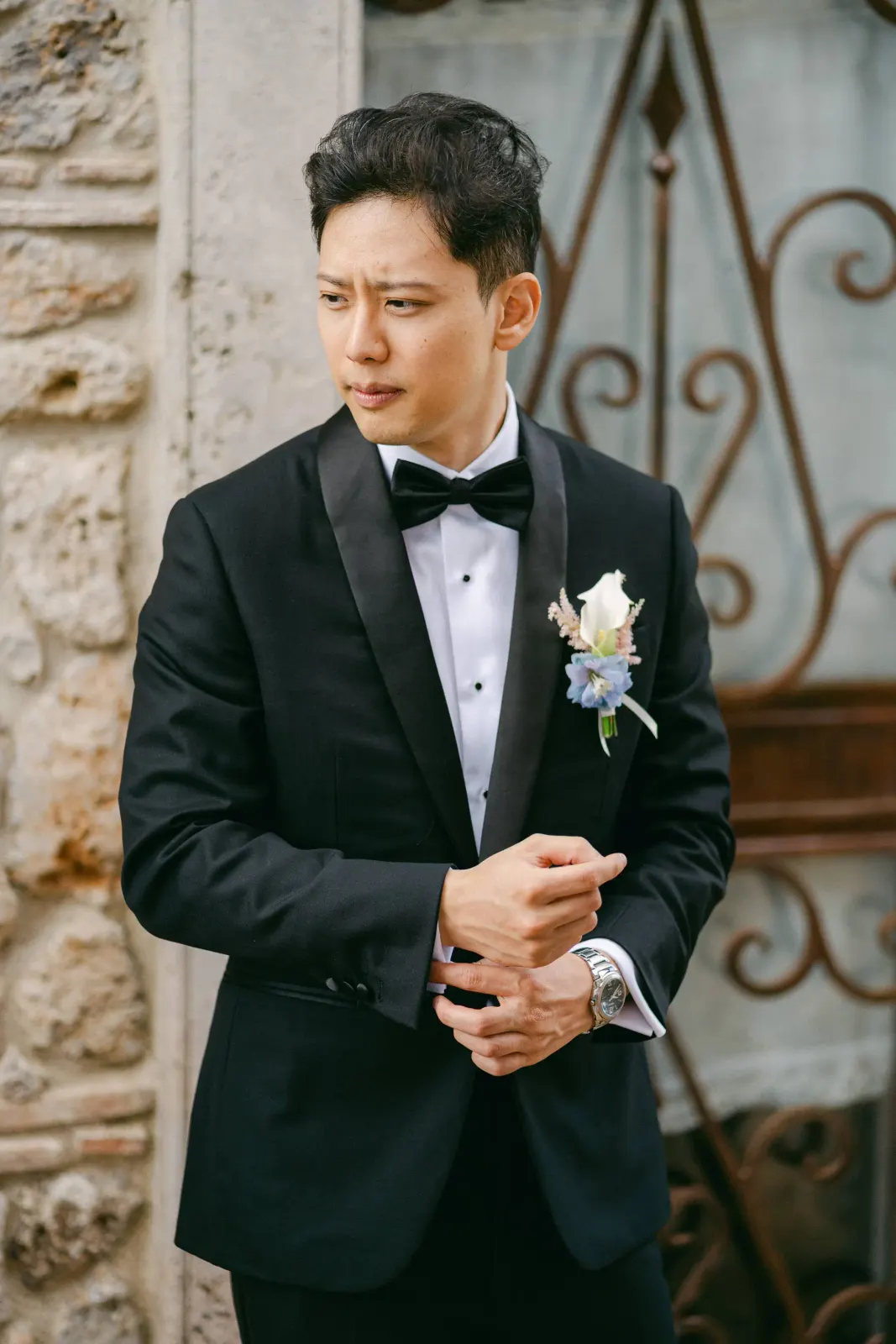 James in tuxedo and bow tie adjusting his cuff, portrait against the stone of the villa