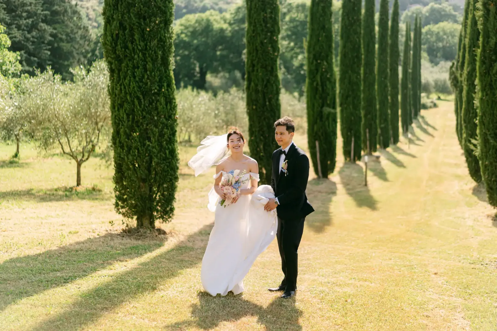 The couple laughing in the cypress avenue with the veil lifted by the wind