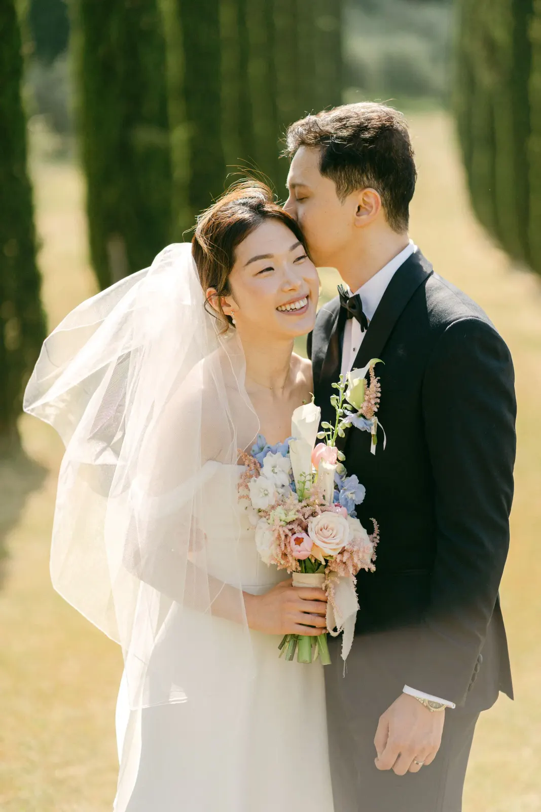 James kissing Audrey's temple as she laughs, with her veil and bouquet