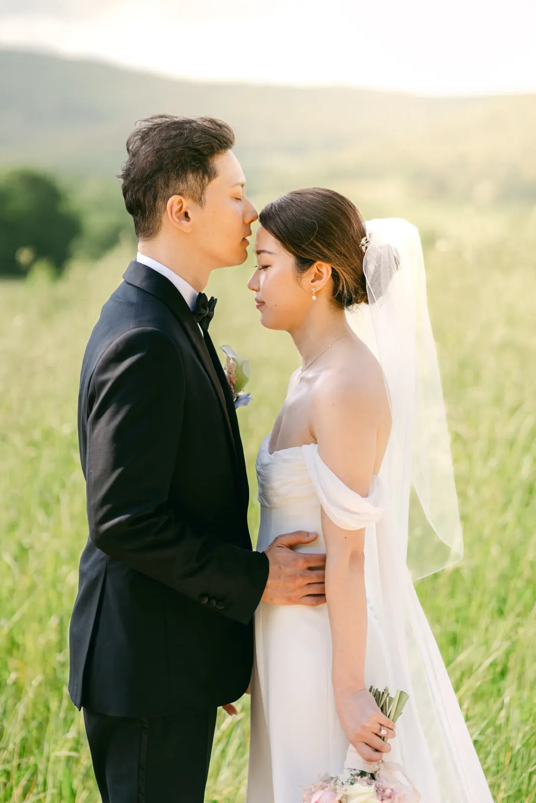 James kissing Audrey's forehead in a field of tall grass at sunset
