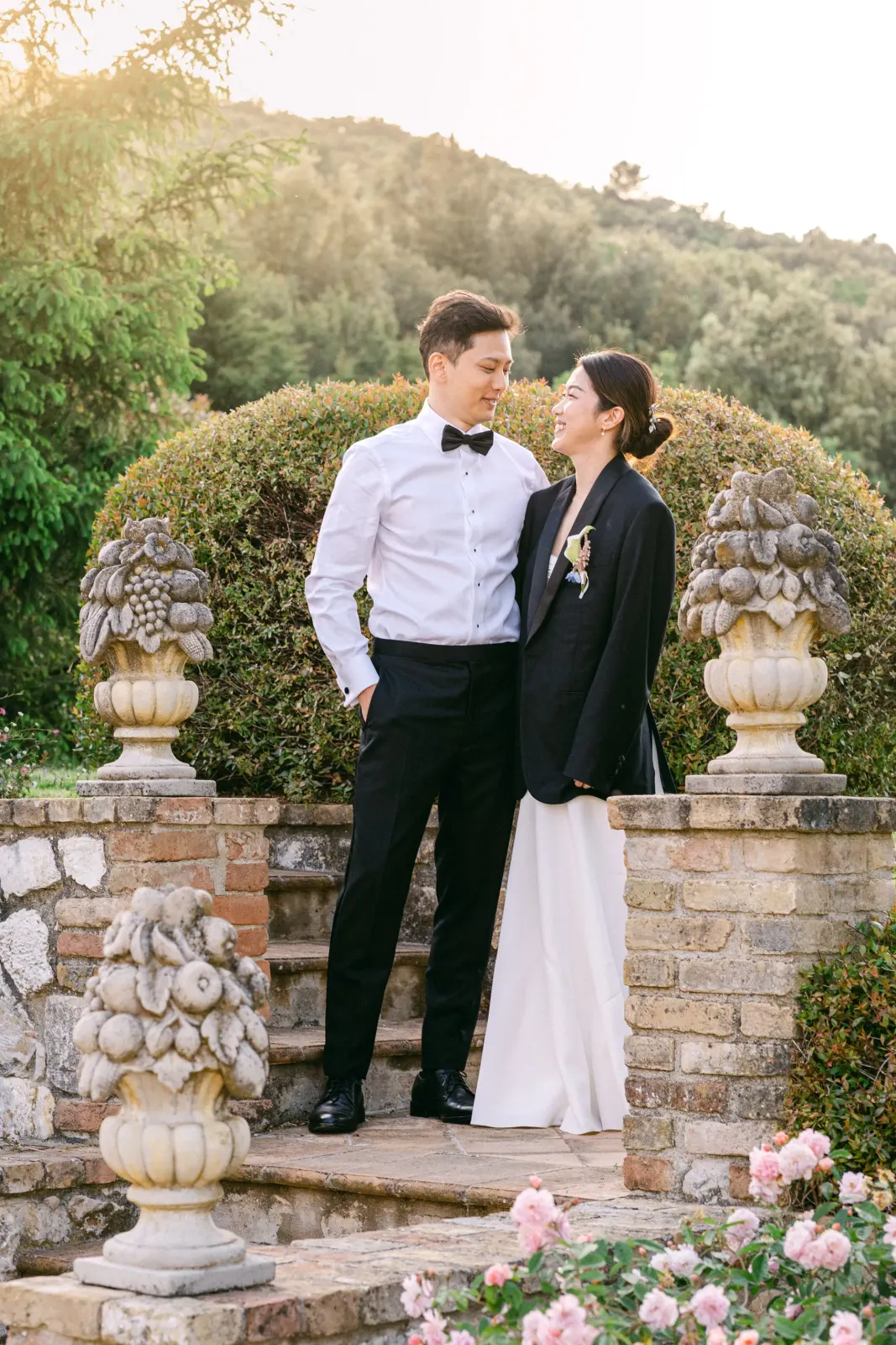The couple on the garden steps between the stone urns, the bride in a black jacket