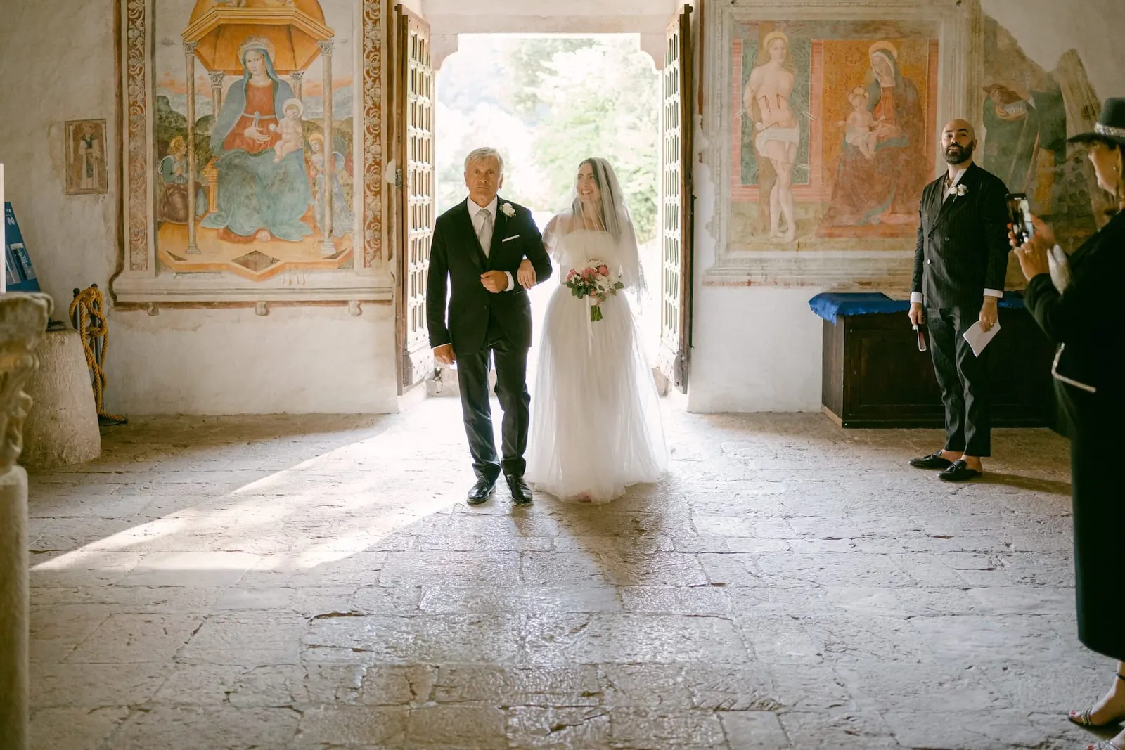 The bride and her father entering the church in backlight, among the frescoes