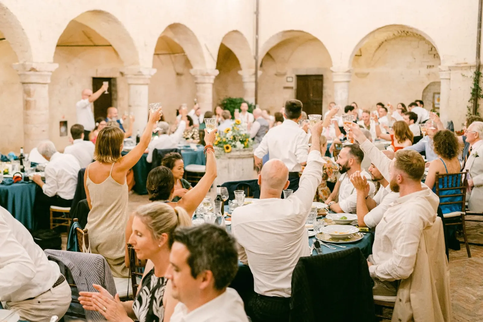 Gli invitati esultano a braccia alzate ai tavoli nel chiostro