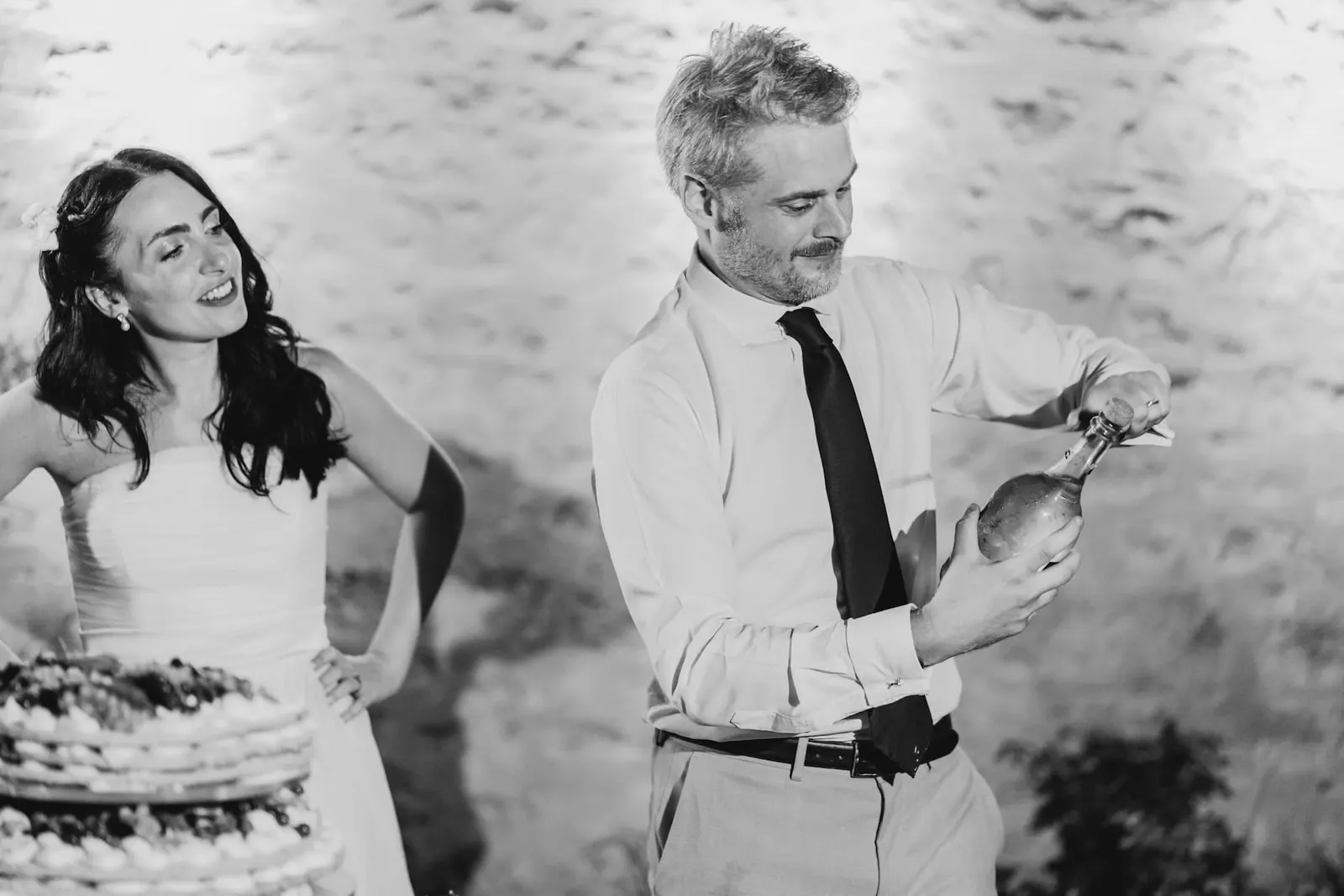 Thomas stappa lo spumante accanto alla torta mentre Valentina lo guarda, in bianco e nero