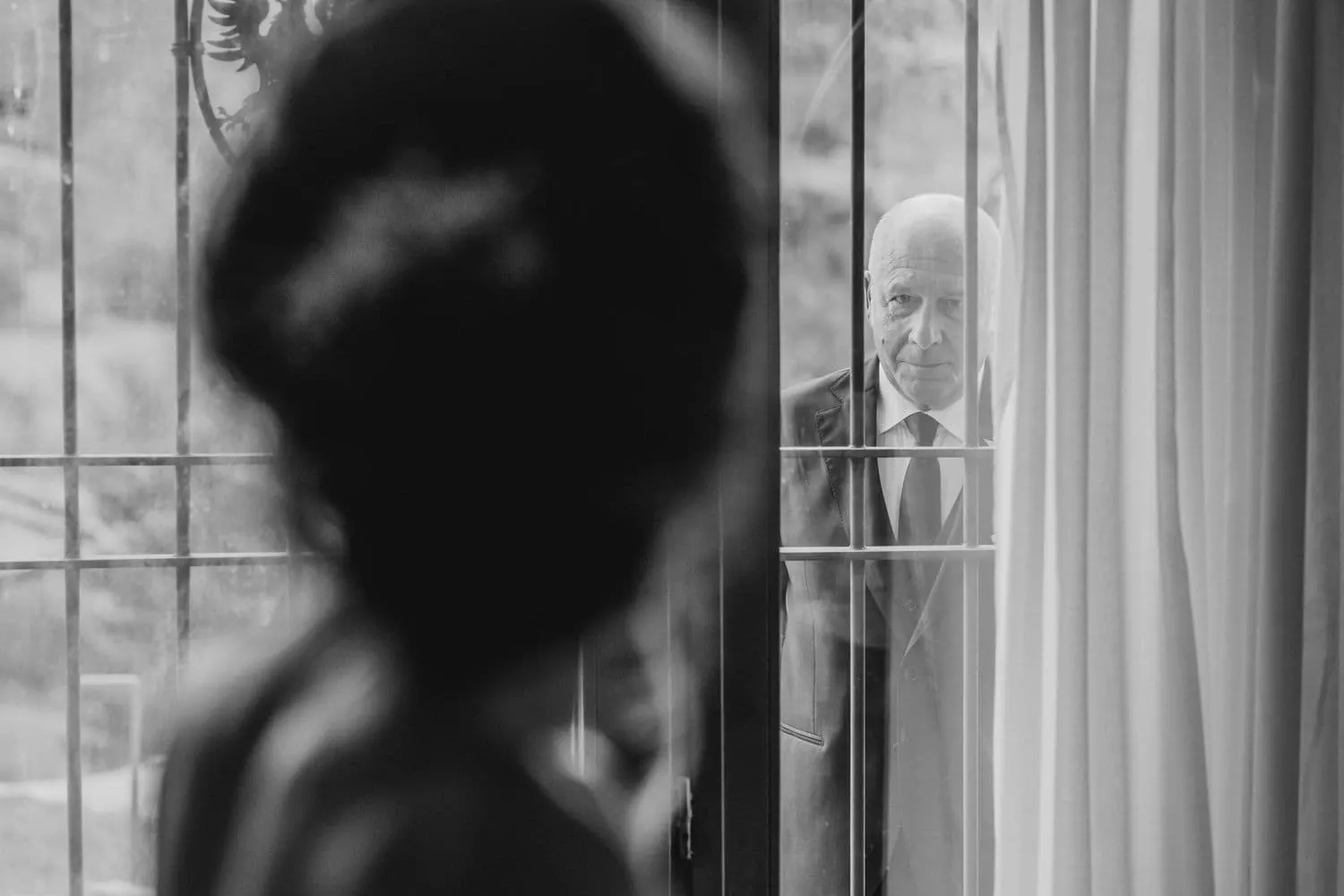 The bride seen from behind in silhouette and a man waiting for her beyond the glass door, in black and white