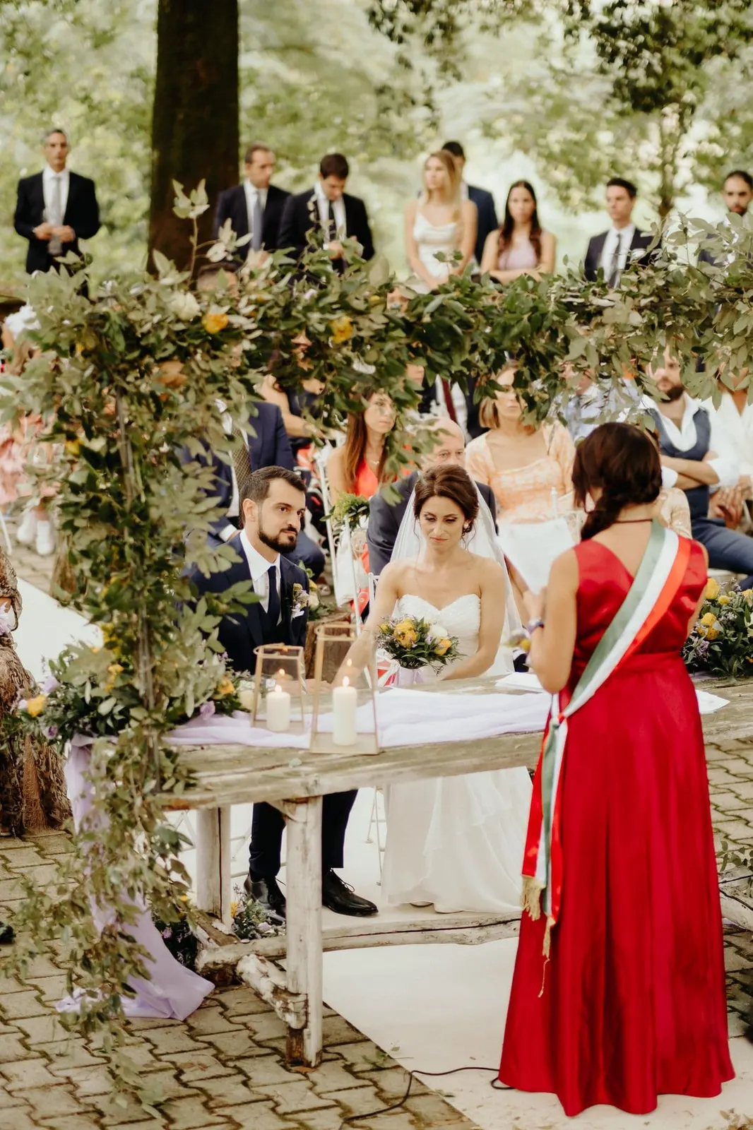 The couple seated at the ceremony table under the leafy arch, in front of the officiant