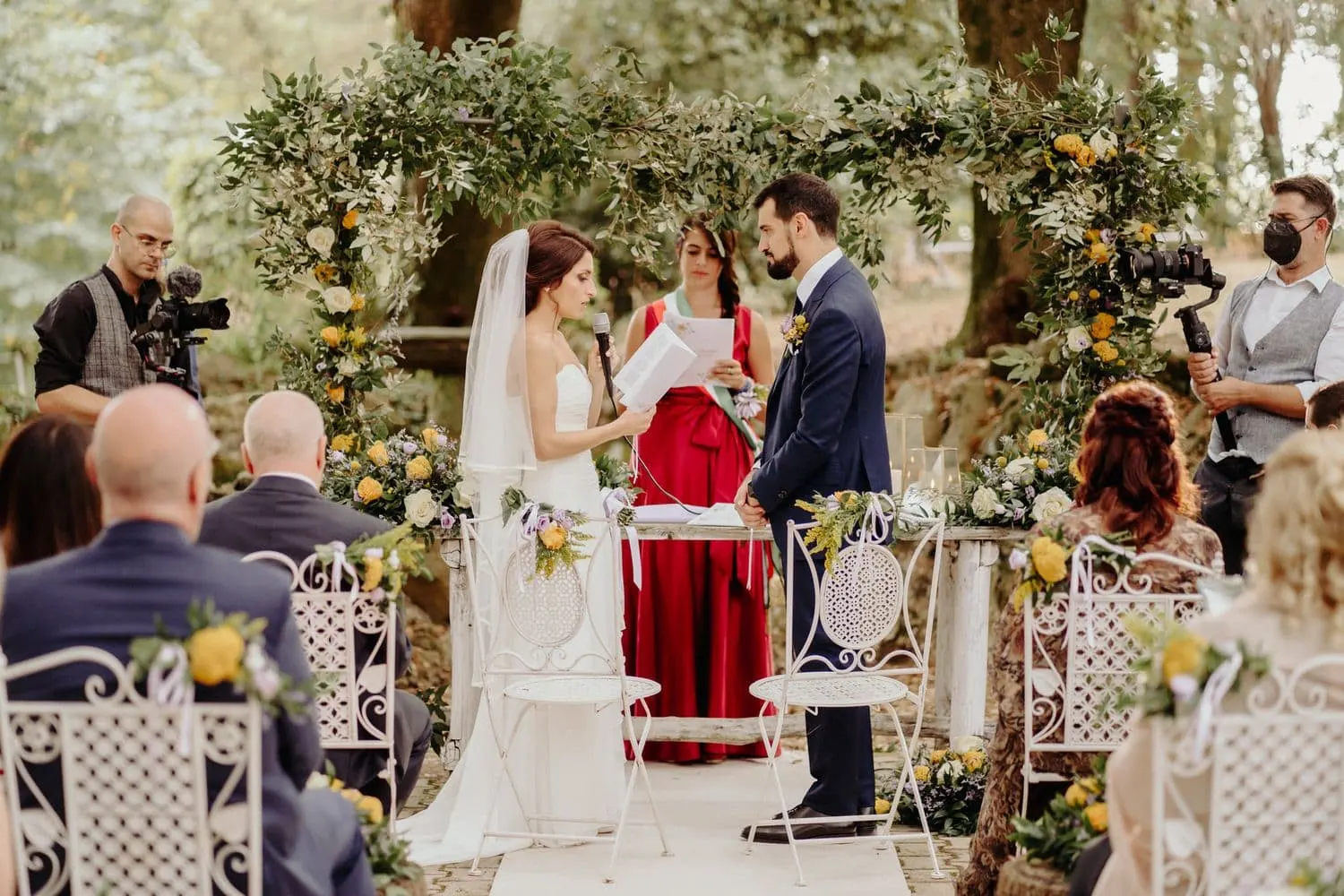 The couple standing before the officiant under the green arch, among the guests