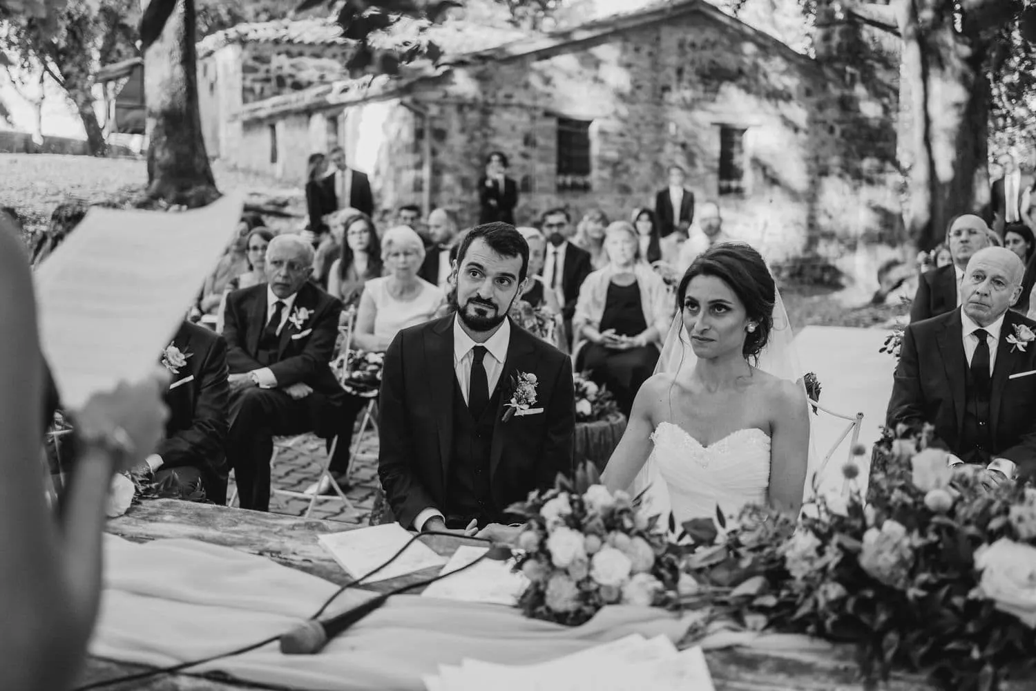 The couple seated during the ceremony, with the guests behind them, in black and white