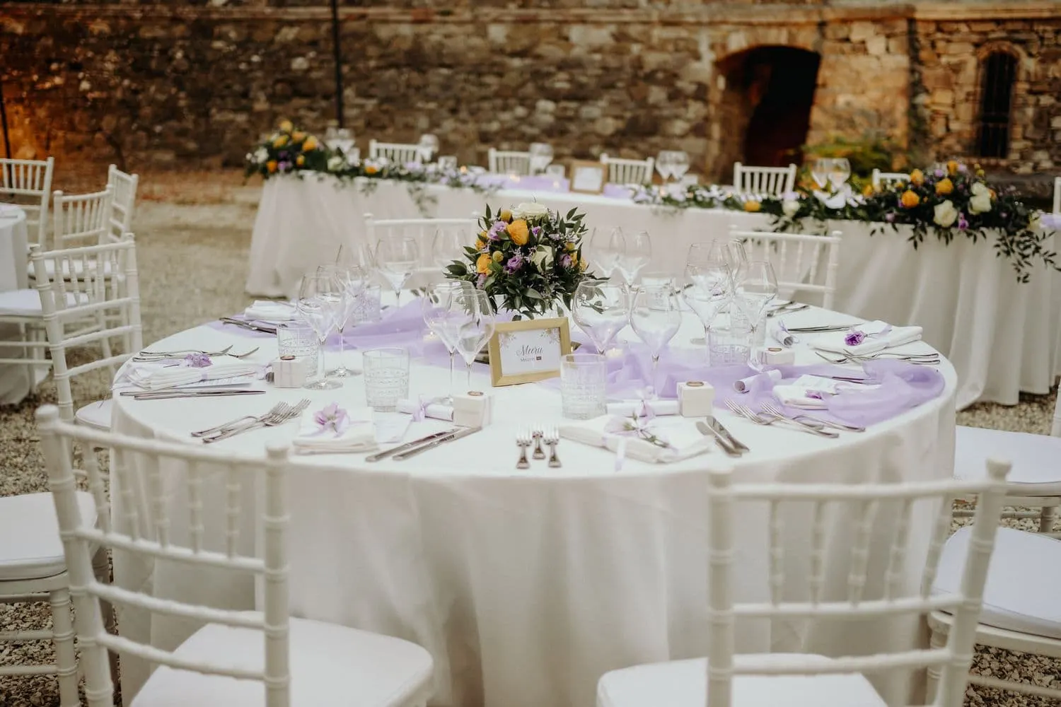 A round table laid outdoors with flowers and lilac details, in front of the stone wall